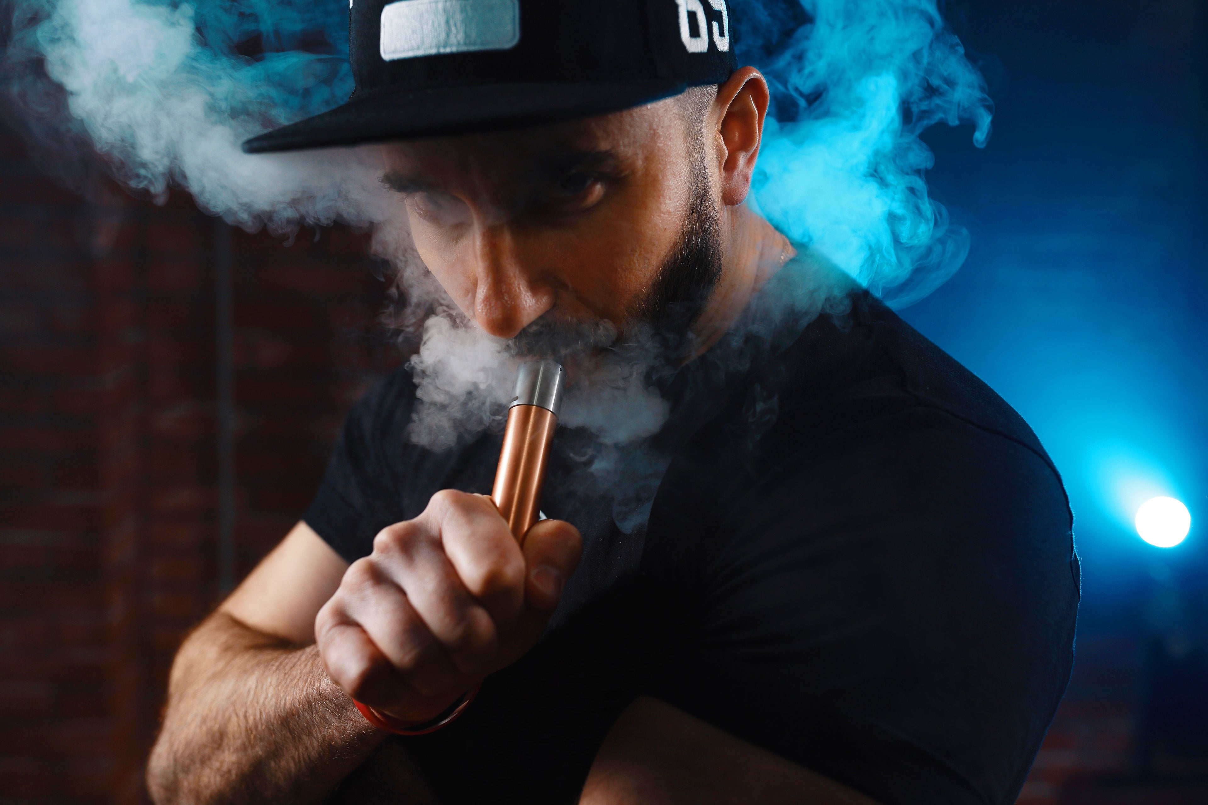 A man wearing a hat with a cigarette in his mouth, looking contemplative against a blurred background.
